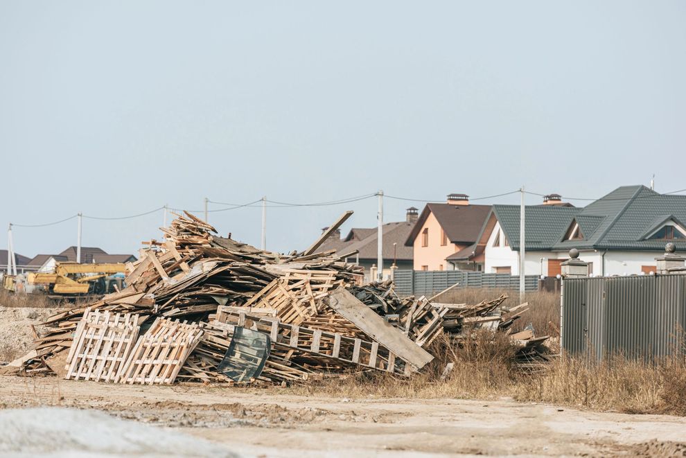 Un grande e disordinato cumulo di pallet di legno usati per il trasporto merci giace su un terreno sterrato di fronte ad alcune case di periferia.