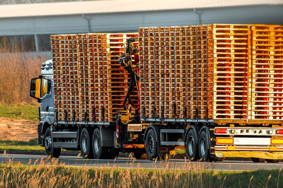 Un camion a pianale ribassato carico di numerose pile di pallet di legno per spedizioni percorre una strada al tramonto.