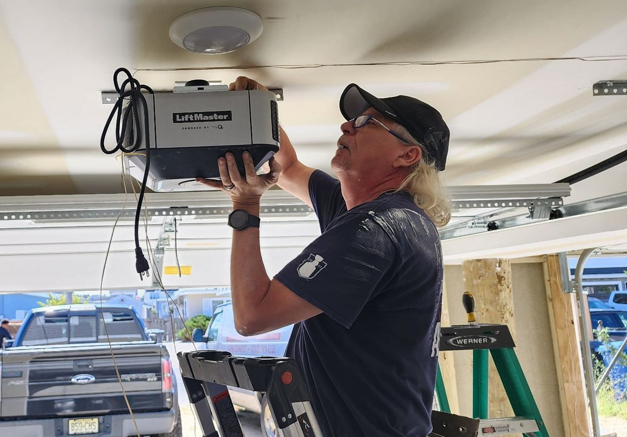A person standing on a ladder installing a LiftMaster garage door opener unit to a garage ceiling.