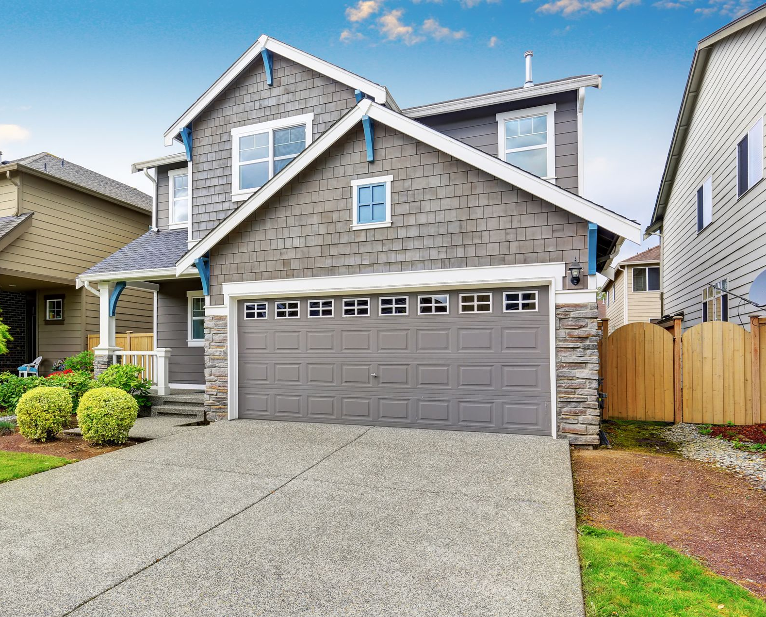A gray multi-story house with a double garage, front yard landscaping, and a concrete driveway under a blue sky.