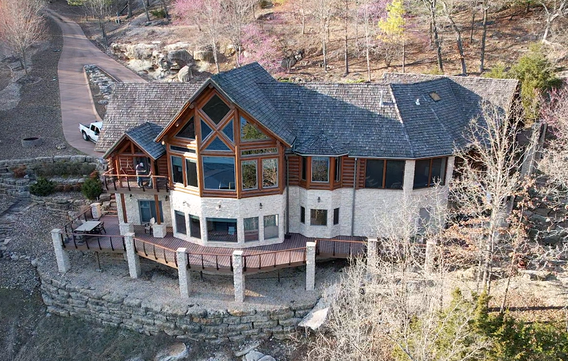 A large stone and wood lakeside house with a steep roof, viewed from above among bare trees.