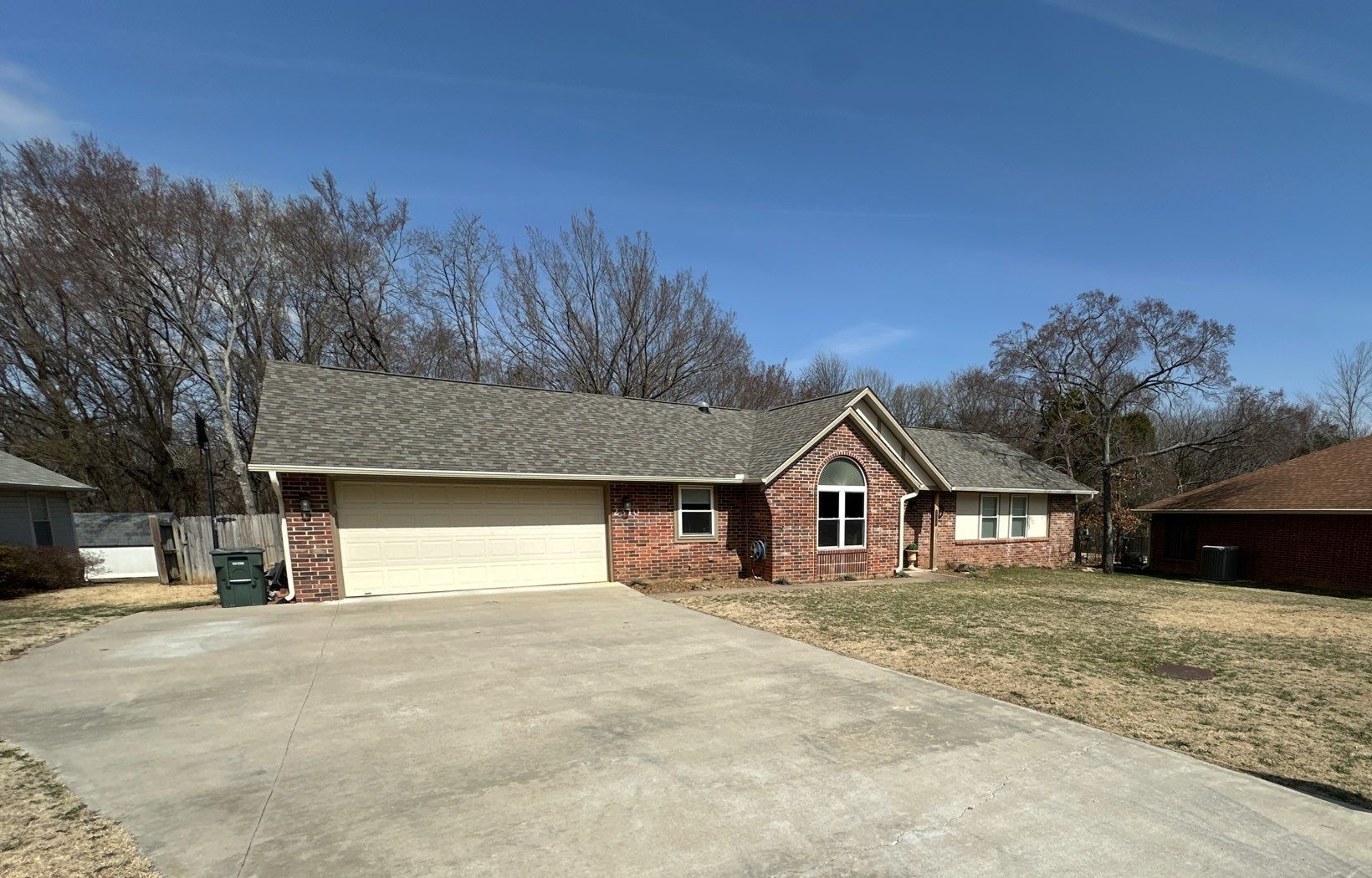 Brick single-story house with beige garage, front driveway, and leafless trees under a clear blue sky