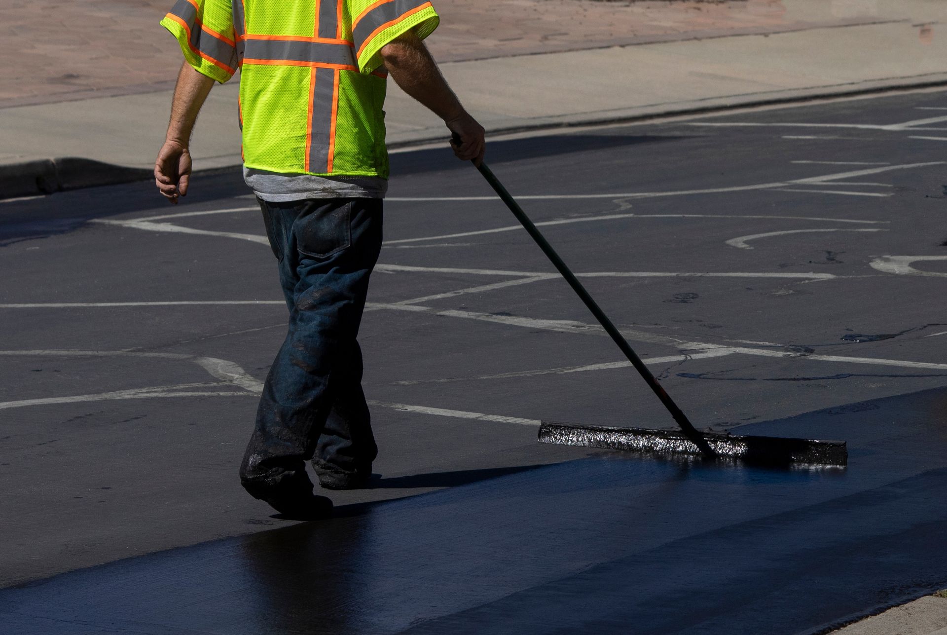 Worker in a reflective vest spreading black pavement coating with a long squeegee.