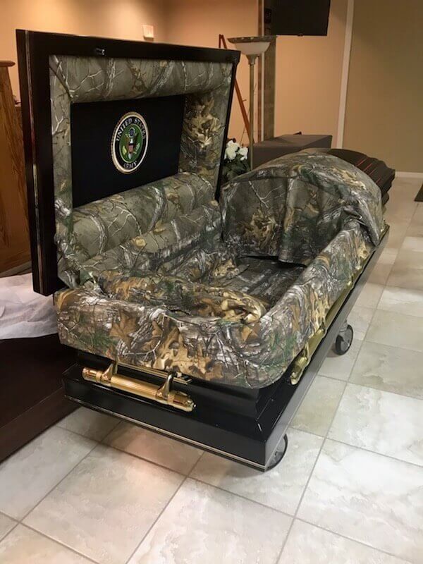Open casket with camouflage lining and emblem. Black exterior, gold handles, in a building.