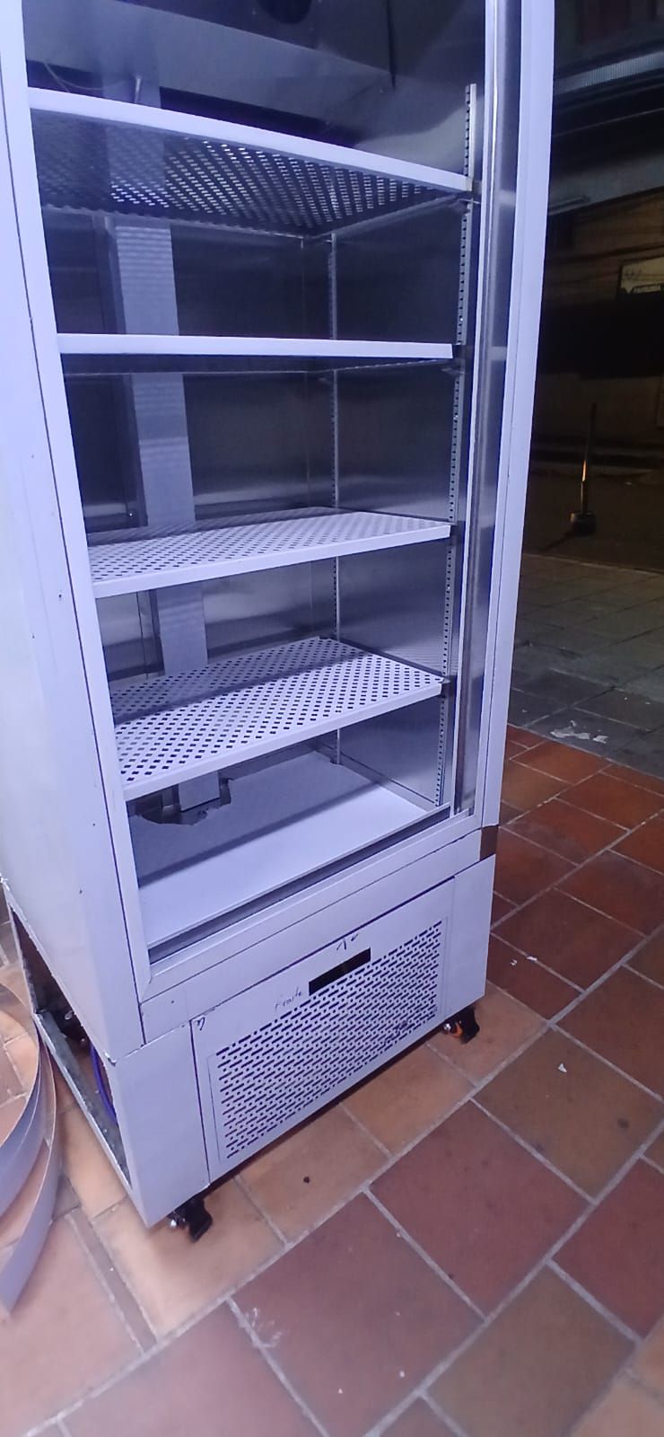Refrigerador alto con puerta de cristal sobre un suelo de baldosas cerca de una pared por la noche.