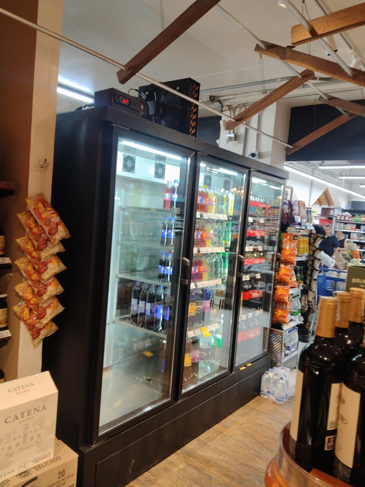 Pasillo de supermercado con refrigeradores con puertas de vidrio, botellas de vino y exhibidores colgantes de aperitivos.
