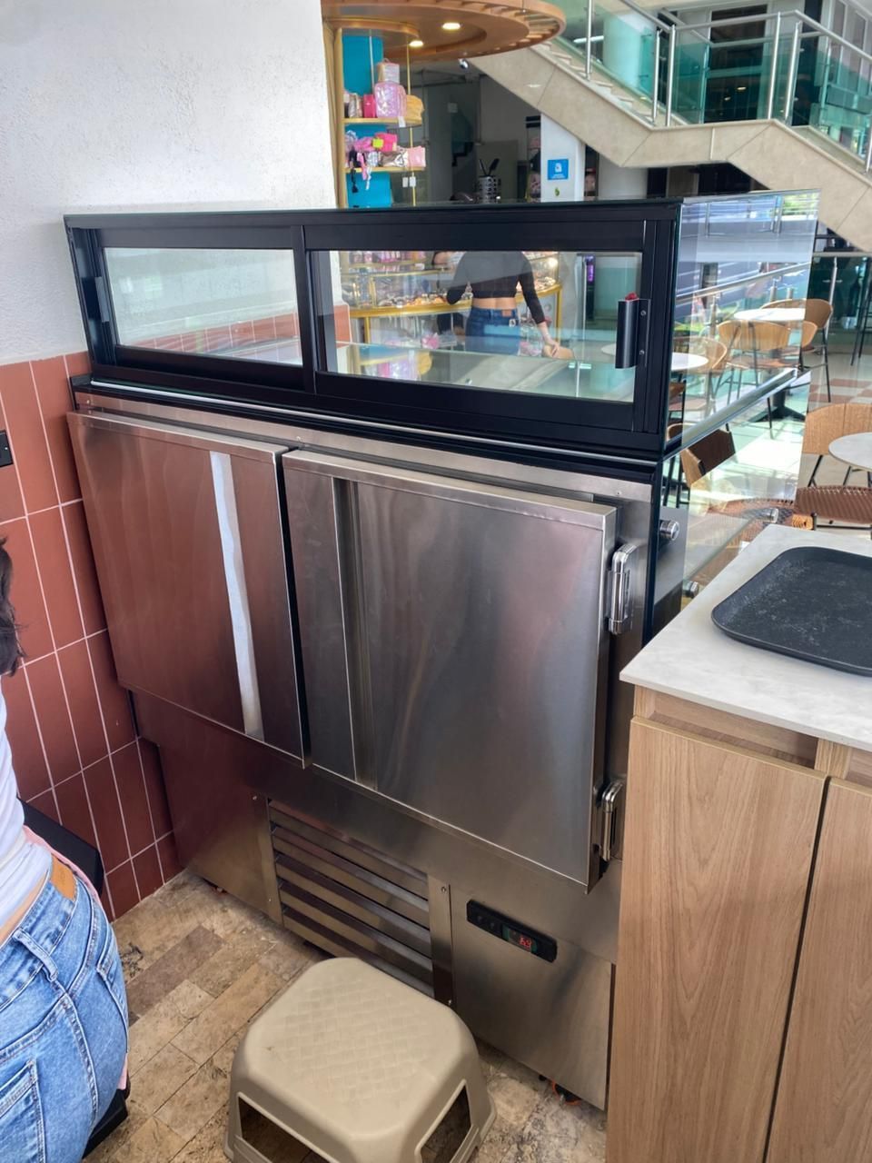 Refrigerador de preparación de acero inoxidable para cafetería con vitrina de cristal y pequeño taburete al lado.