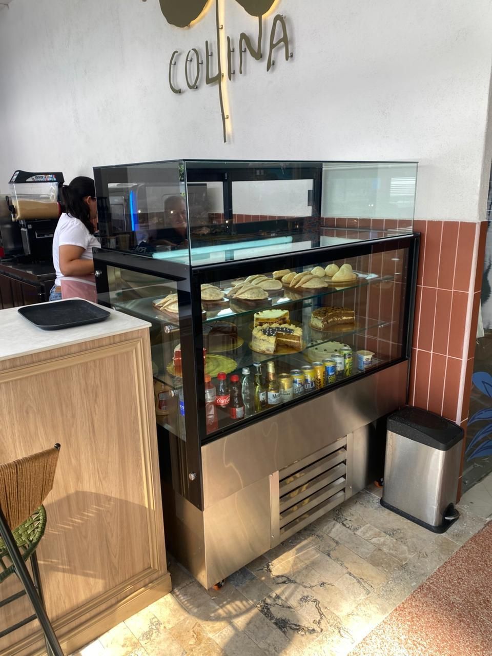 Vitrina de cristal con pasteles en su interior y una persona de pie detrás del mostrador en una cafetería.
