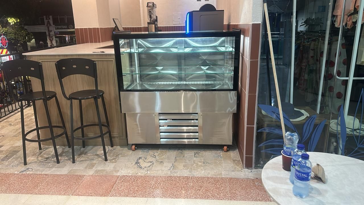Pequeña barra de cafetería con vitrina refrigerada y dos taburetes en el interior.
