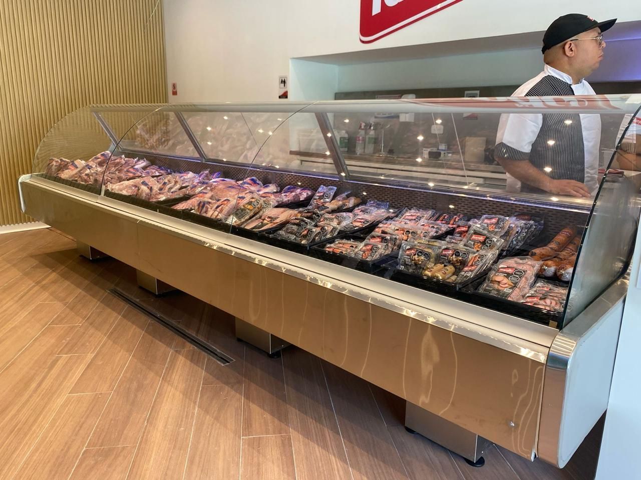 Vitrina larga para carne en una tienda, con un empleado detrás del mostrador y suelo con diseño de madera.