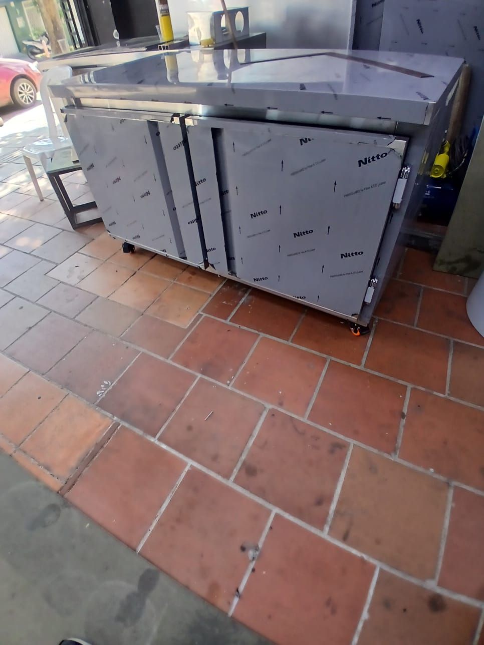 Caja de almacenamiento de metal gris en un patio embaldosado junto a un edificio, con un coche visible al fondo.