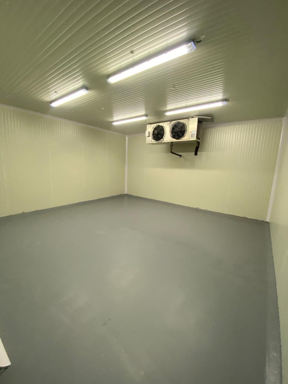Cámara frigorífica vacía con paredes blancas, suelo gris, luces fluorescentes y ventiladores montados en la pared.