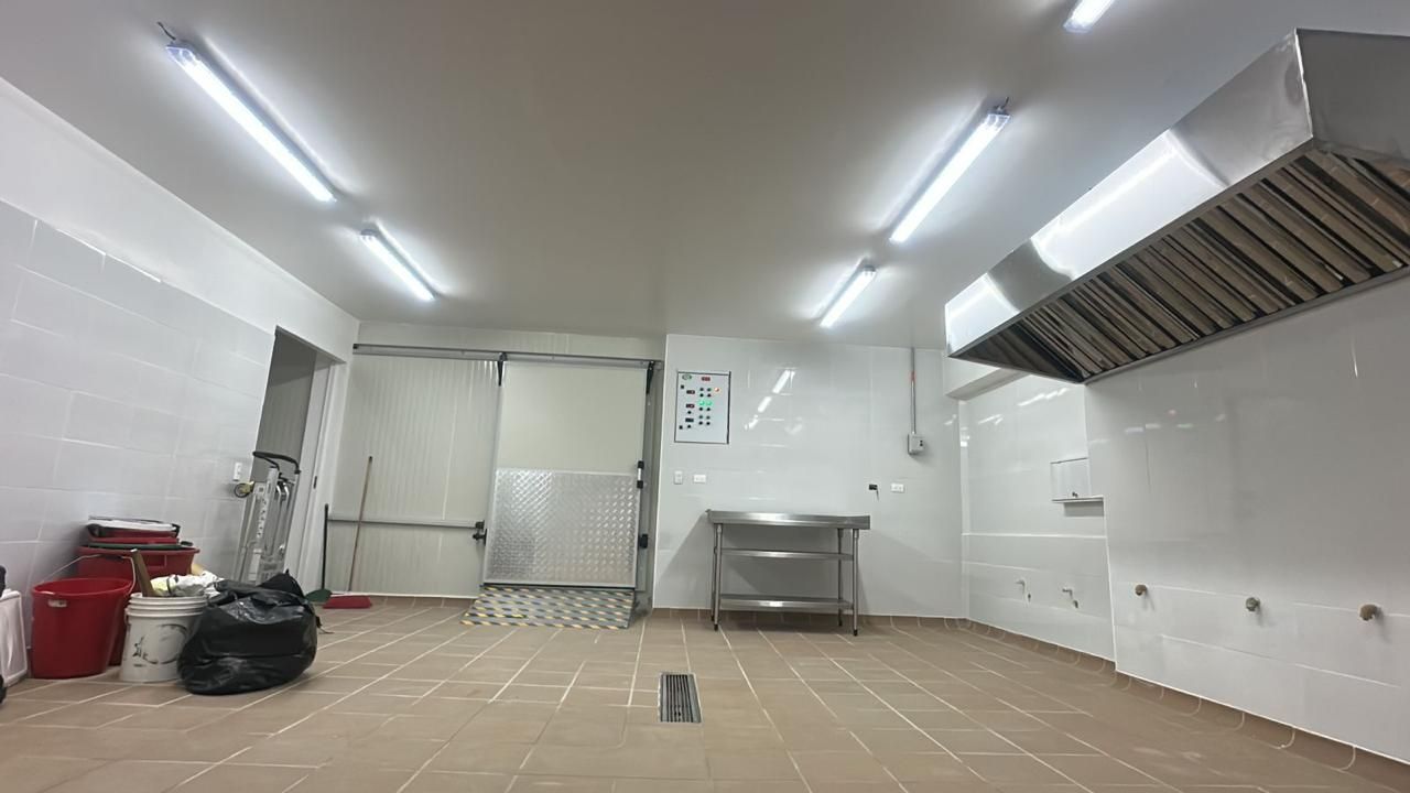 Habitación vacía con azulejos blancos, luces fluorescentes, mesa de metal, lavabo y productos de limpieza.