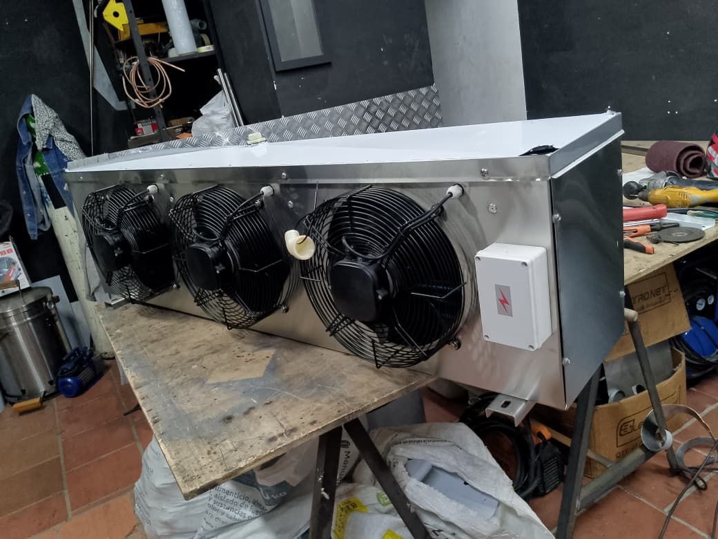 Caja metálica casera con tres ventiladores negros sobre un banco de trabajo desordenado en un taller.