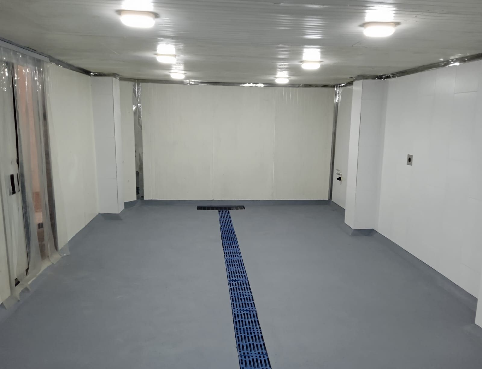 Habitación blanca vacía con suelo gris y una franja azul en el centro bajo las luces del techo.