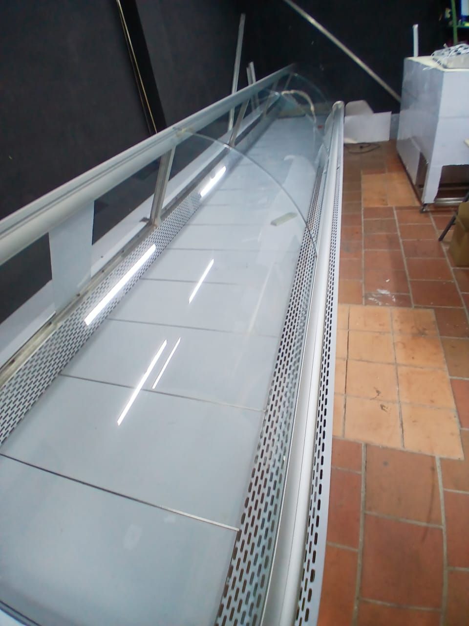 Cinta transportadora metálica o vitrina expositora de alimentos con tapa de cristal sobre suelo de baldosas en interiores.