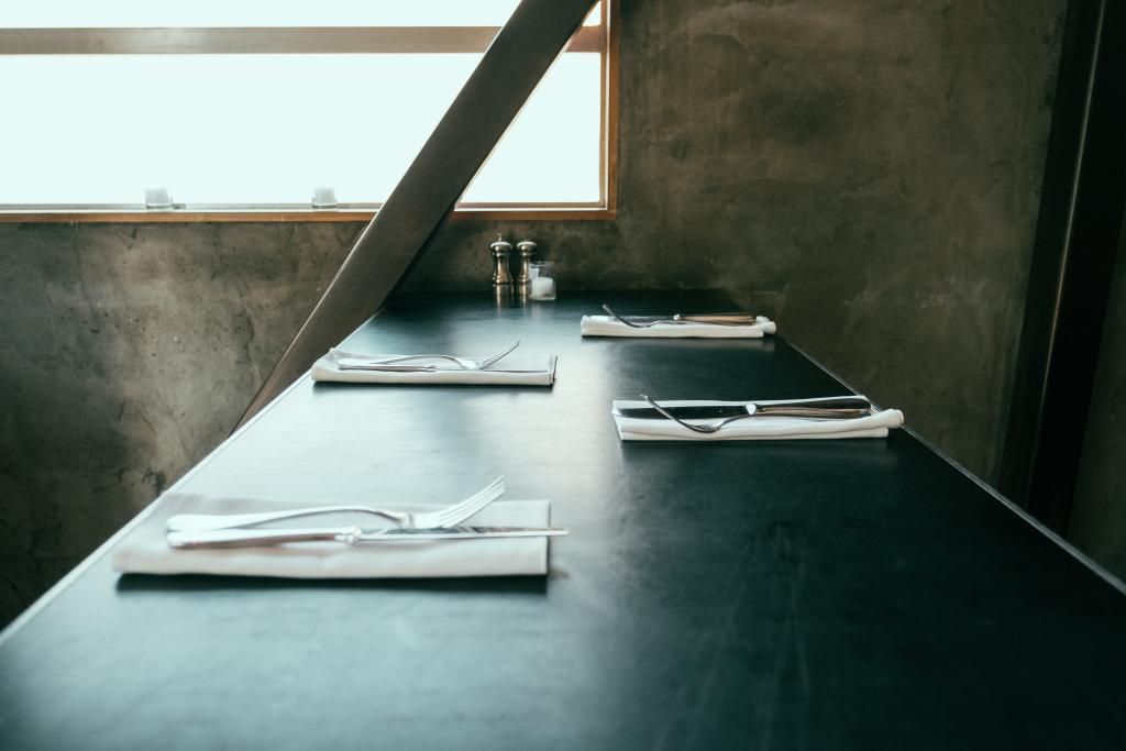 Mesa de comedor minimalista oscura junto a una ventana, con platos y cubiertos blancos en una habitación moderna.