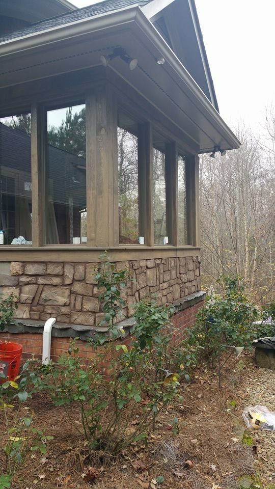 Stone-sided house porch with large windows, overhanging roof, and a wooded yard with sparse plants