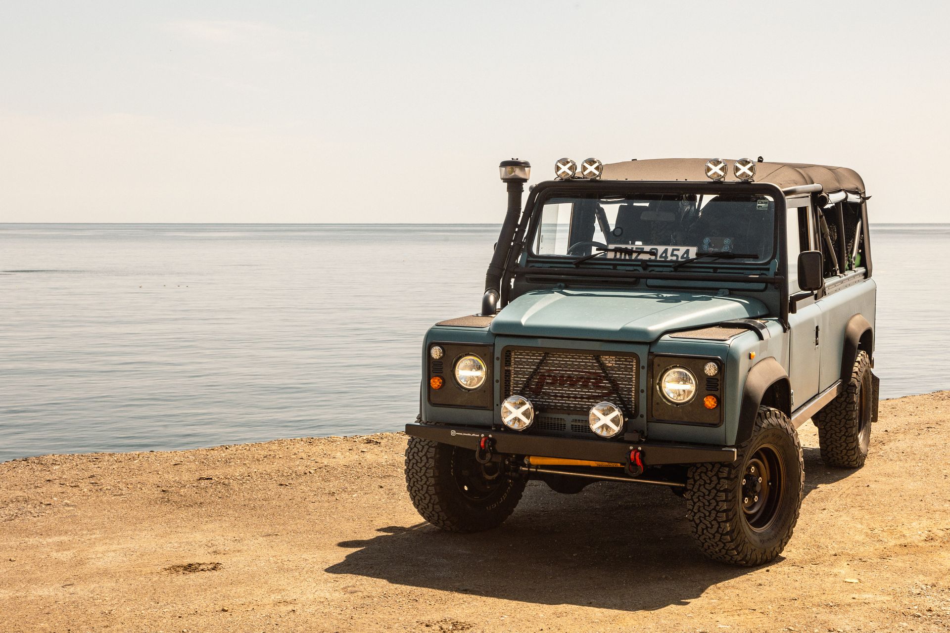 Blue off-road vehicle parked on a sandy beach, facing the ocean on a sunny day.