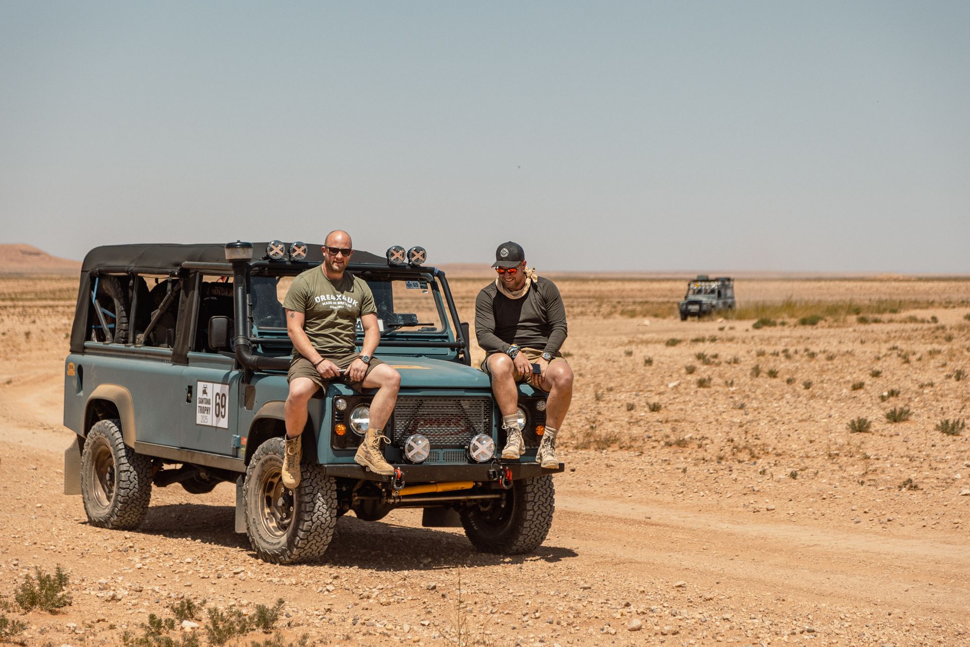Two men sit on the hood of a blue off-road vehicle in a desert. A second vehicle is in the distance.