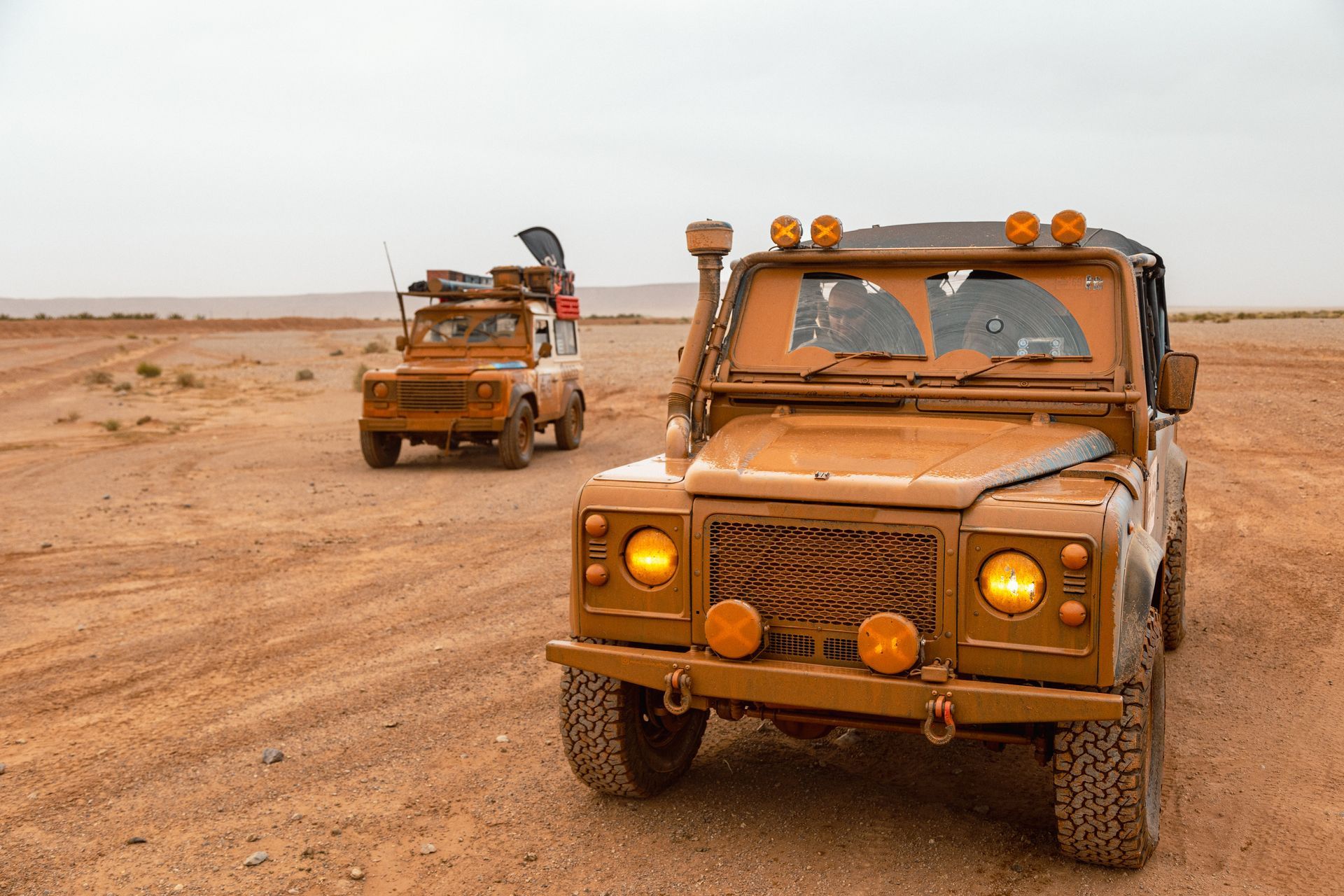 Two mud-covered Land Rover Defenders driving in a desert landscape.