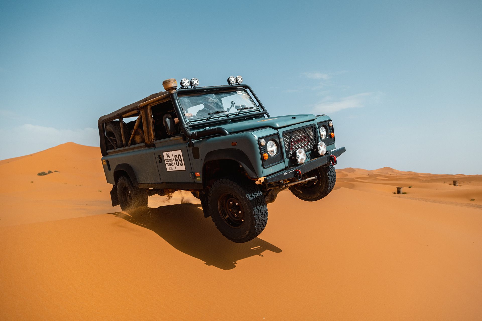 Blue off-road vehicle jumping over a sand dune in a desert setting.