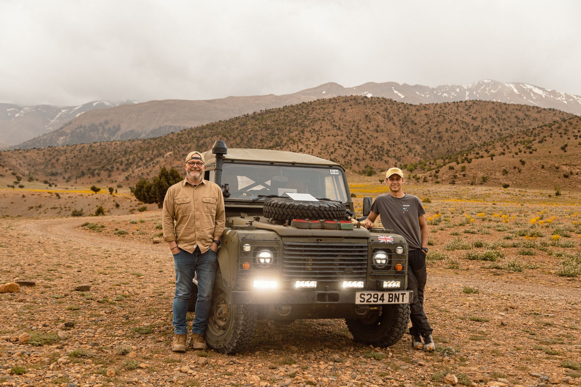 Two men pose beside a green Land Rover in a mountainous desert landscape.