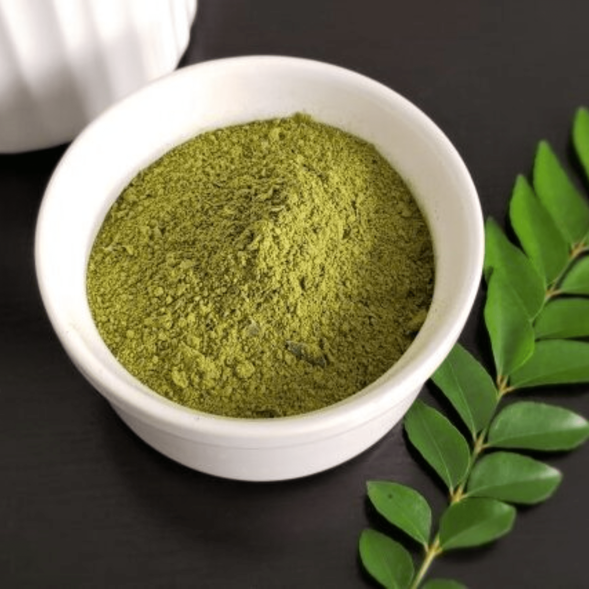 Green curry powder in a white bowl, with fresh curry leaves on a dark surface.
