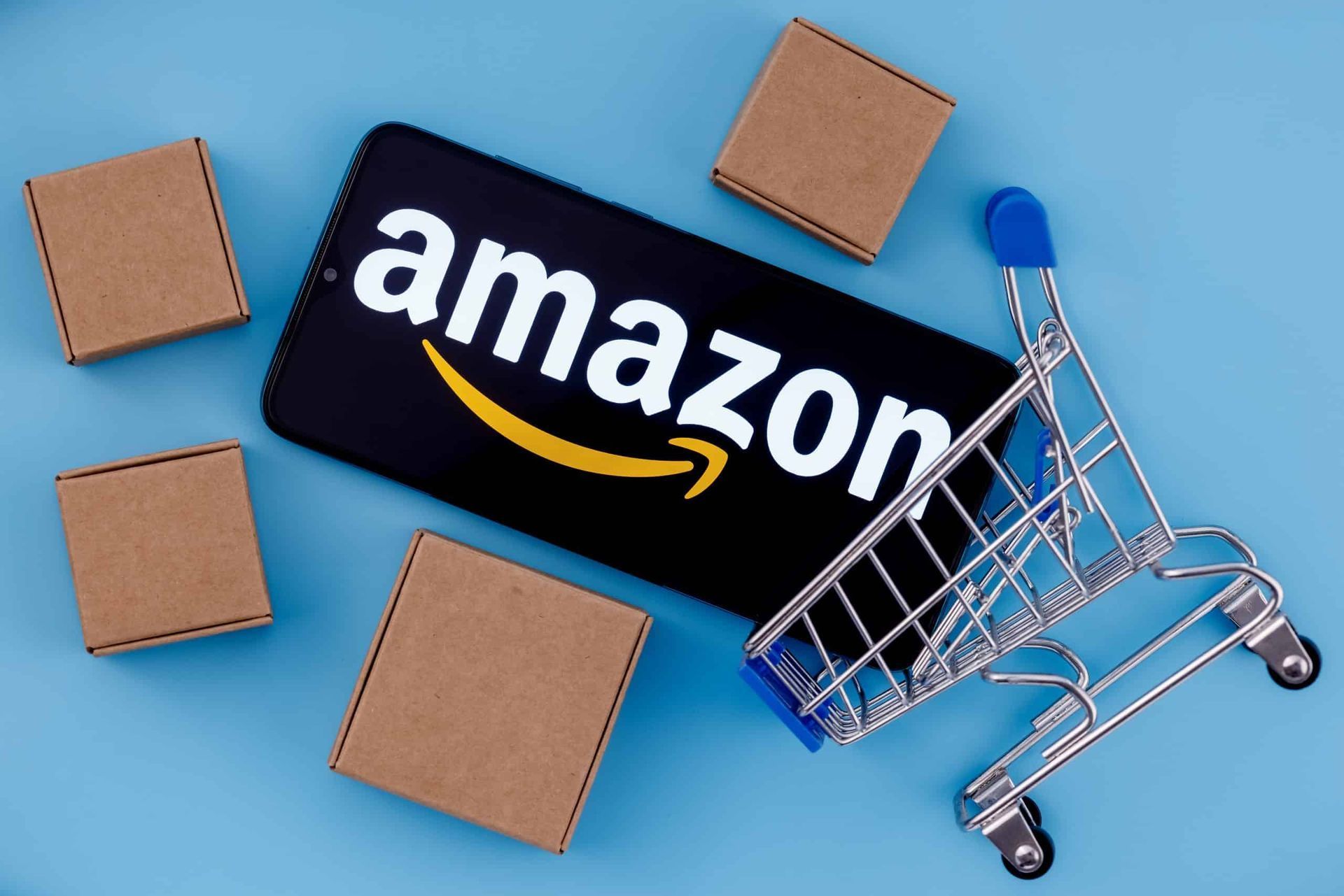 Amazon logo on a phone, small shopping cart, and brown boxes on a blue background.