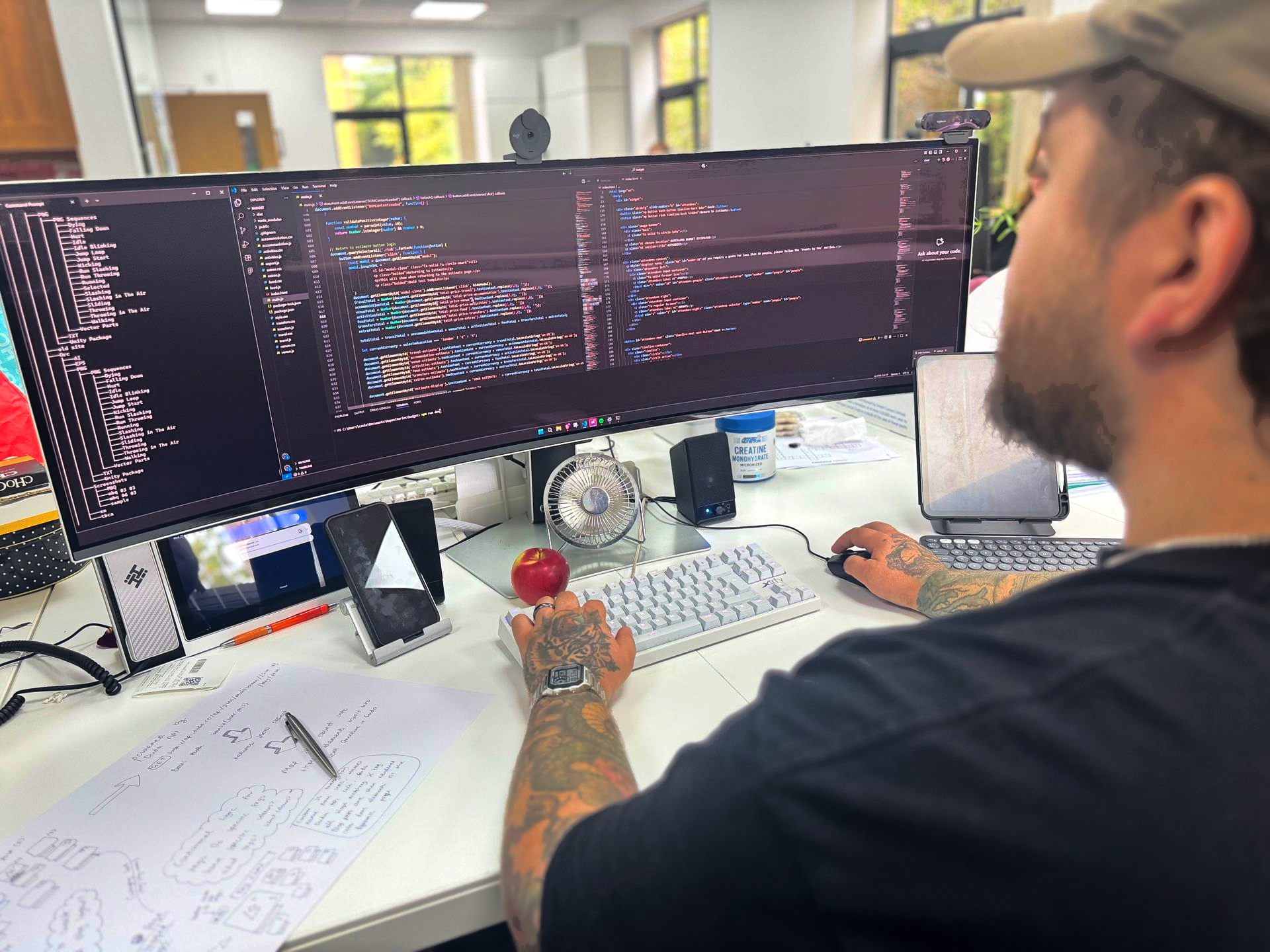 Man with tattoos coding on a multi-monitor setup at a desk, focused gaze, in an office setting.