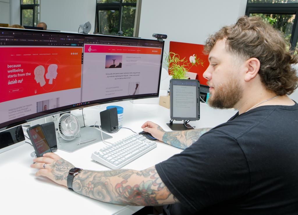 Man with tattoos works at desk with multiple screens and an e-reader.