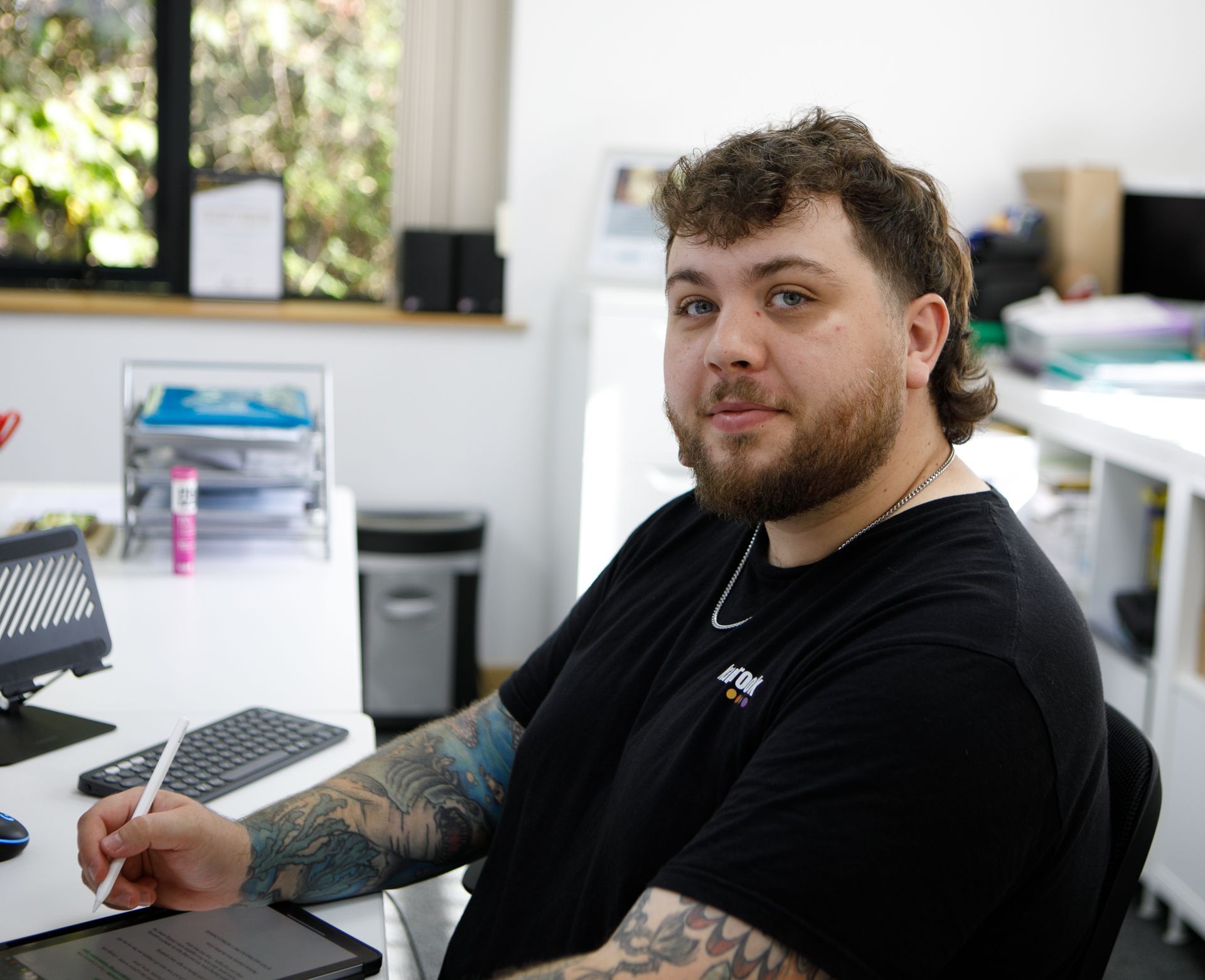 Man with curly hair and tattoos at a desk, smiling while looking at the camera.