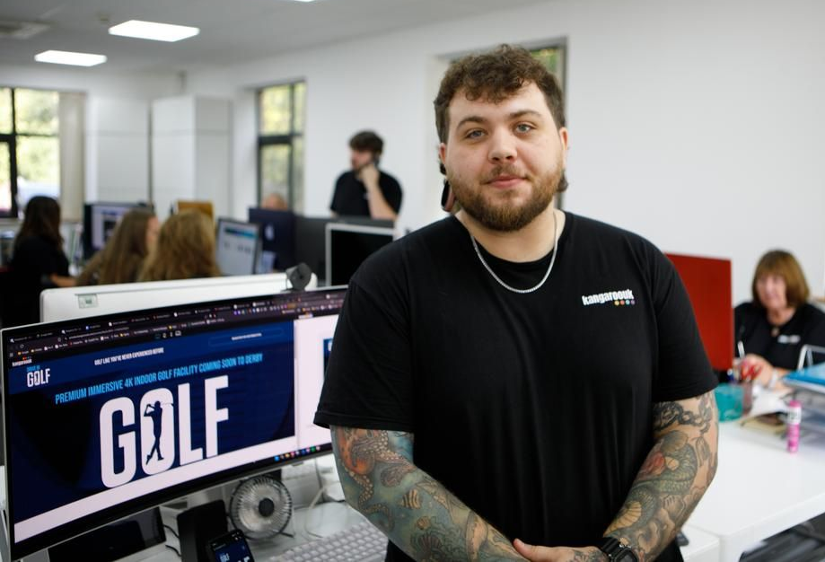 Man with tattoos and a black shirt in an office, standing near a computer screen displaying a golf website.