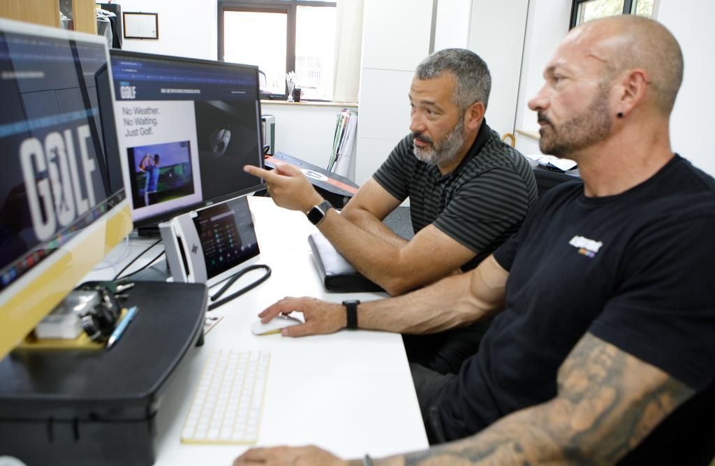 Two men at a computer, one pointing at the screen, discussing online golf content.
