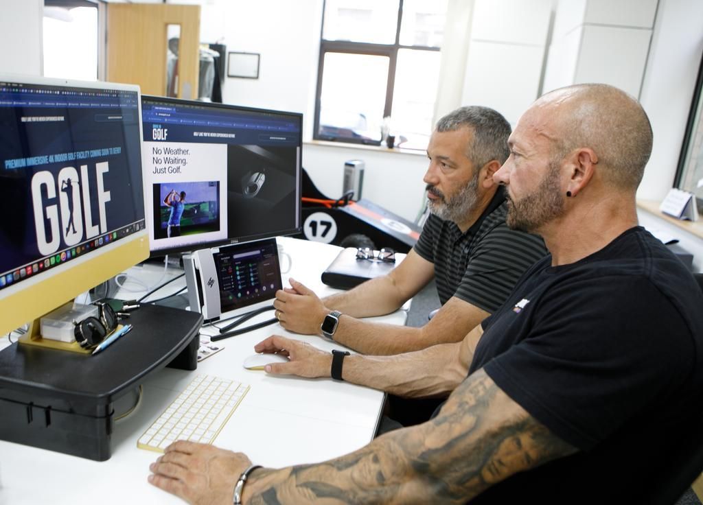 Two men editing on desktop computers in a bright office with multiple monitors showing web pages.