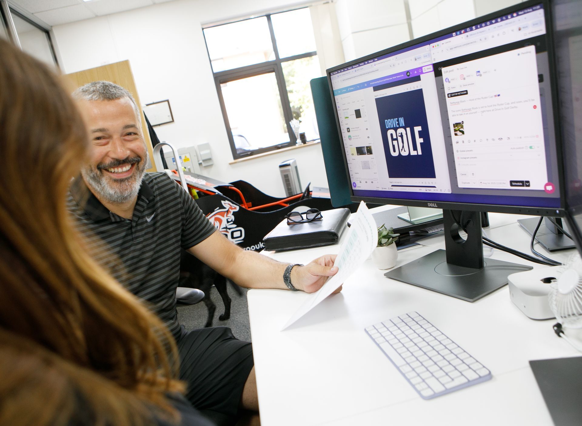 Man smiling, looking at a document with a woman. They are sitting at a desk with two computer monitors.