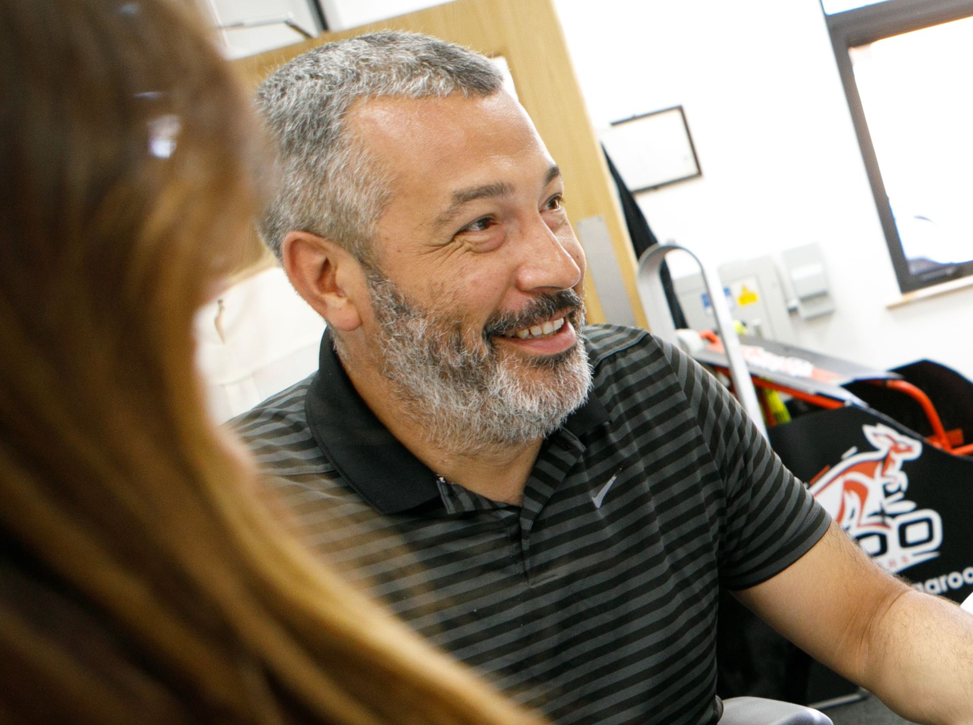 Man with gray beard smiling, wearing a gray striped polo shirt. Sitting indoors, looking at someone off-screen.