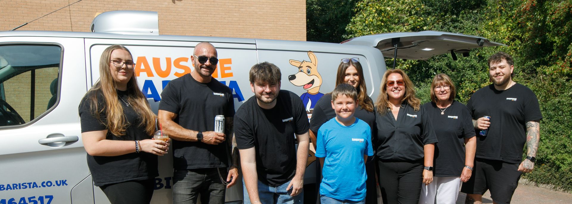Seven people stand smiling in front of a white van with large blue and orange lettering.