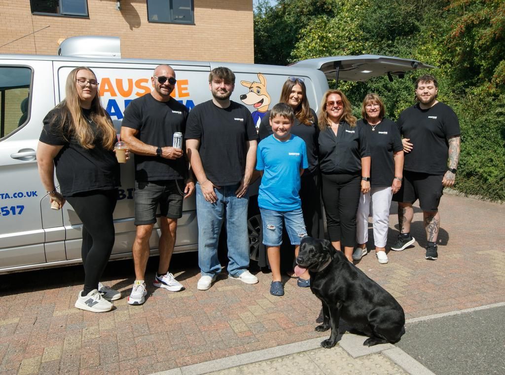 A group of people and a black dog pose in front of a van, under a sunny sky.