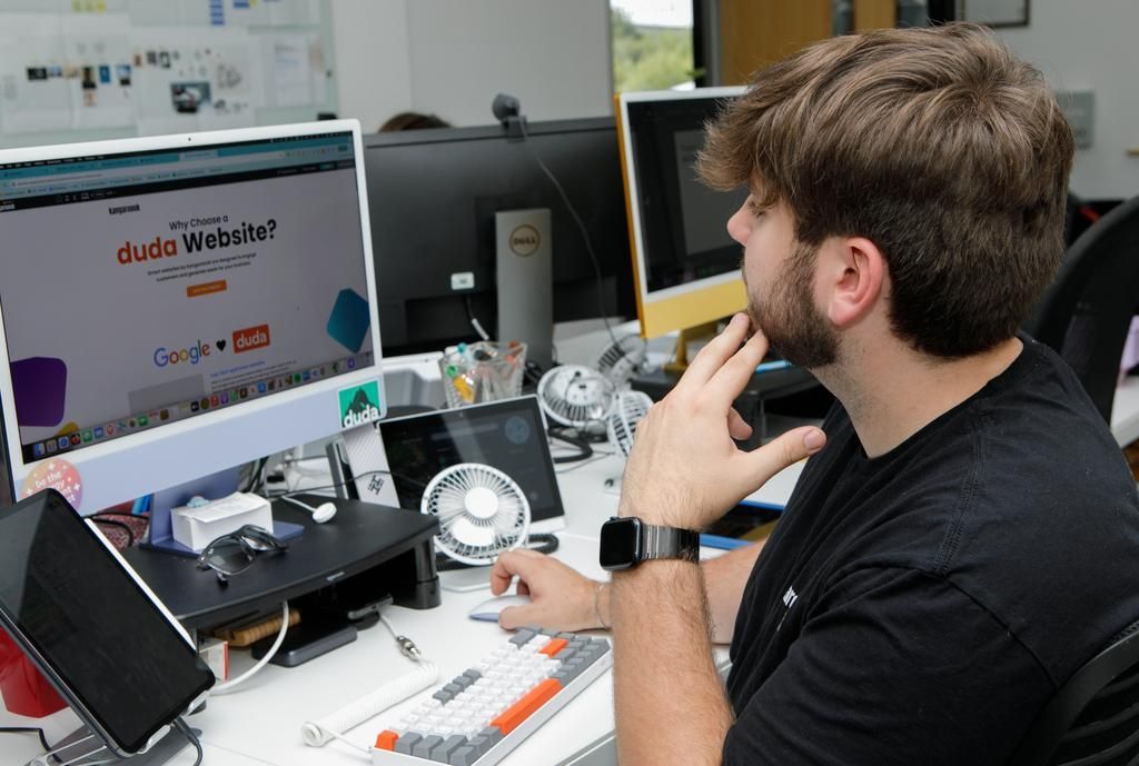 Person at a desk working at dual monitors in an office, holding a mouse and looking at the screen.