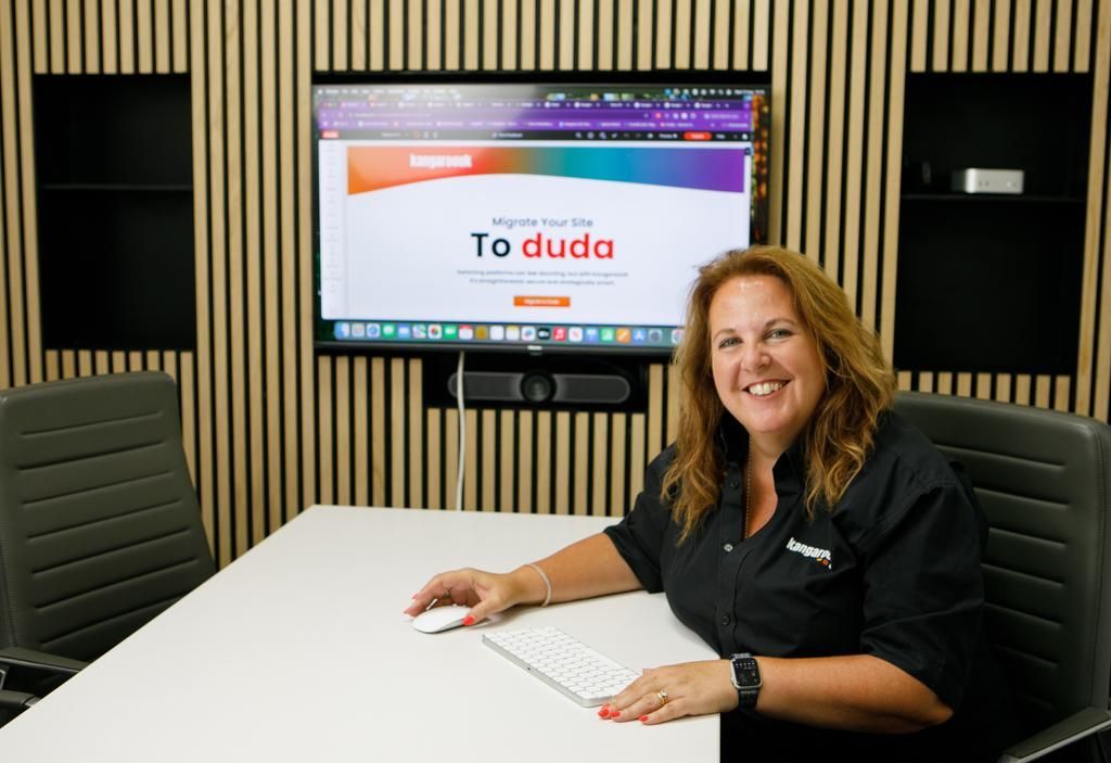 Woman at table in a meeting room; a website is displayed on a screen behind her.