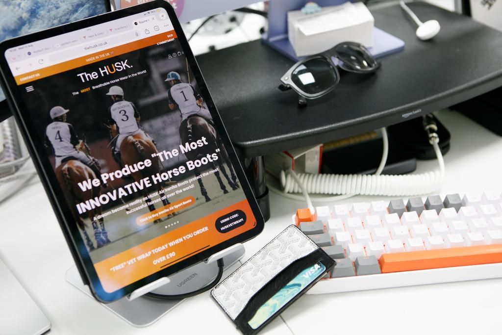 Tablet displaying a website about horse boots; desk setup with keyboard, sunglasses, and wallet.