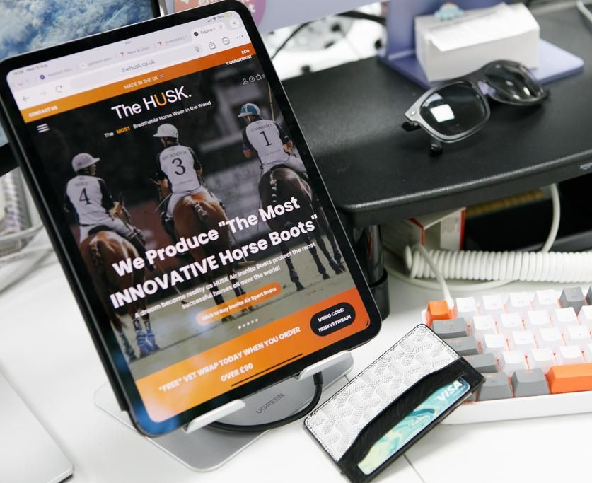 Tablet displaying The HUSK website, with horse riding photo. Sunglasses, keyboard, and wallet also visible.