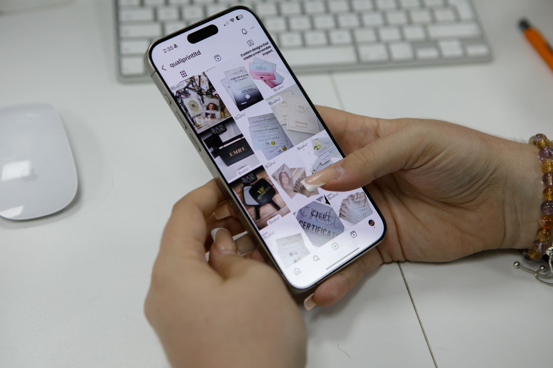 Hands holding a smartphone displaying a grid of product photos on a white desk with a keyboard and mouse.