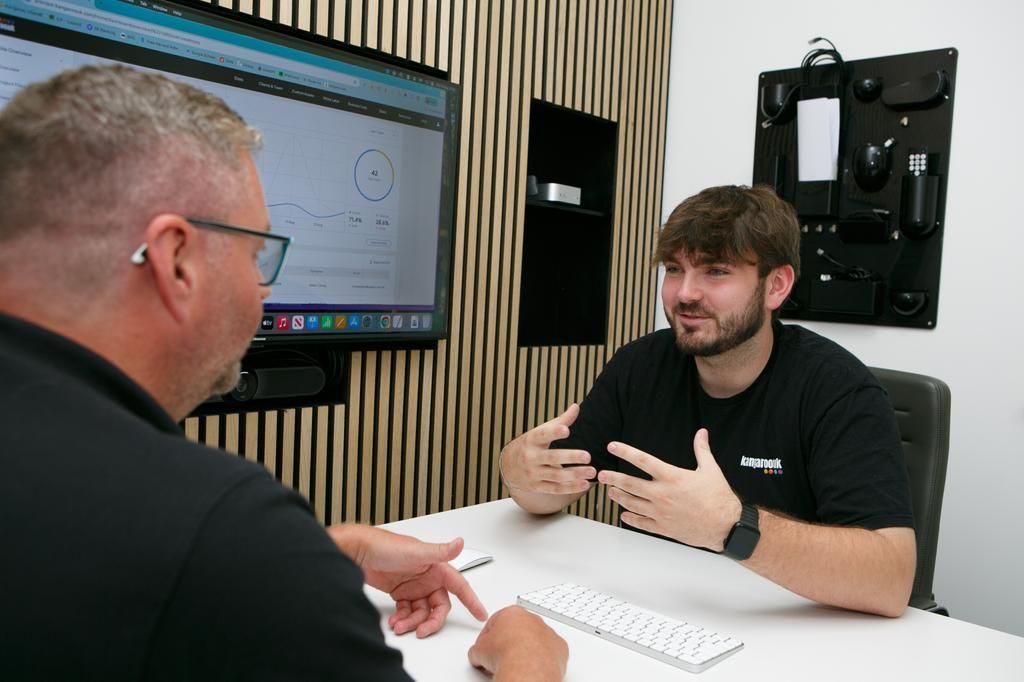 Two men in an office setting, one pointing at a computer screen, the other gesturing.