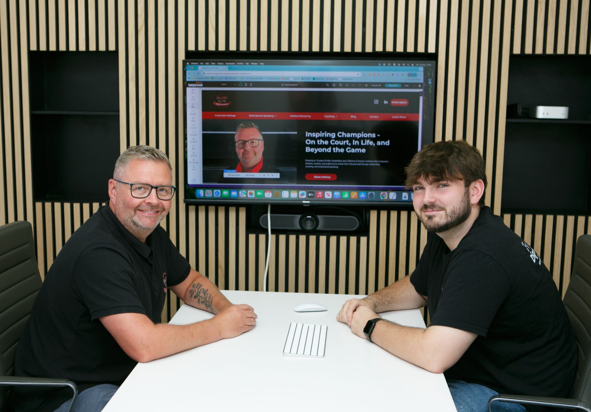 Two people sit at a white desk in an office, facing the camera with a large screen displaying a website in the background.