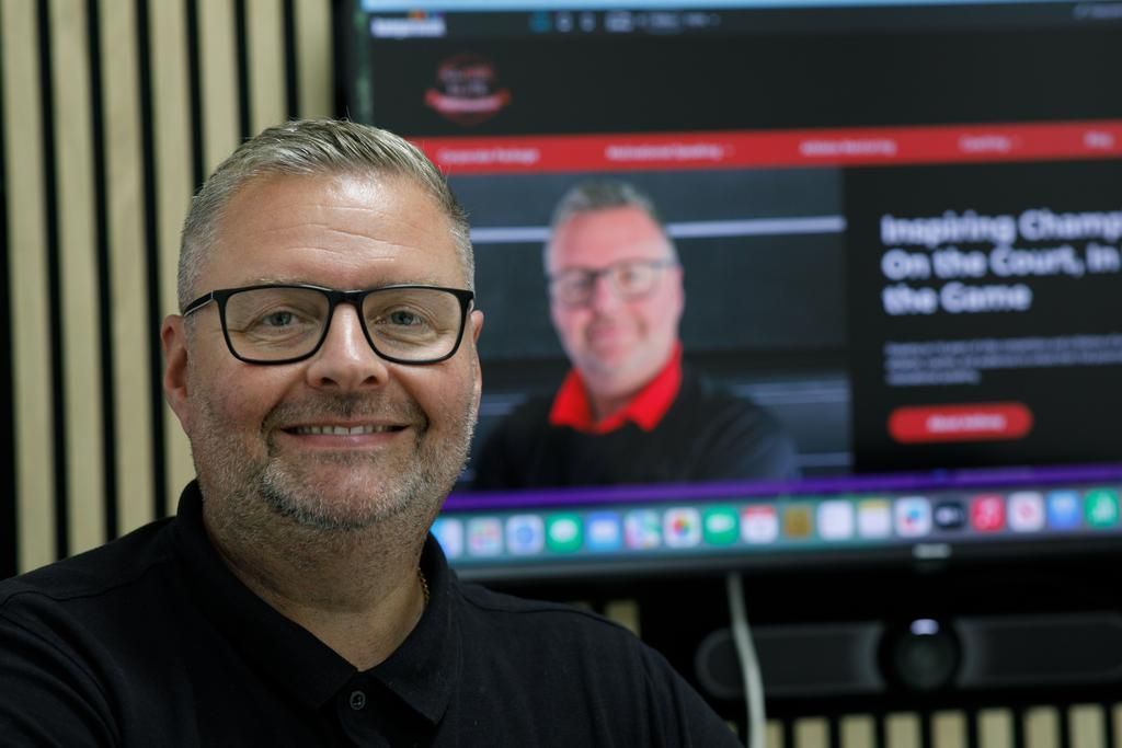 Man with glasses smiling, stands in front of a screen with his image and text,