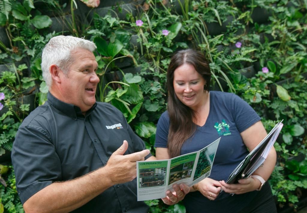 Man and woman reviewing a brochure in front of a green wall. Both smiling, gesturing, and engaged in discussion.