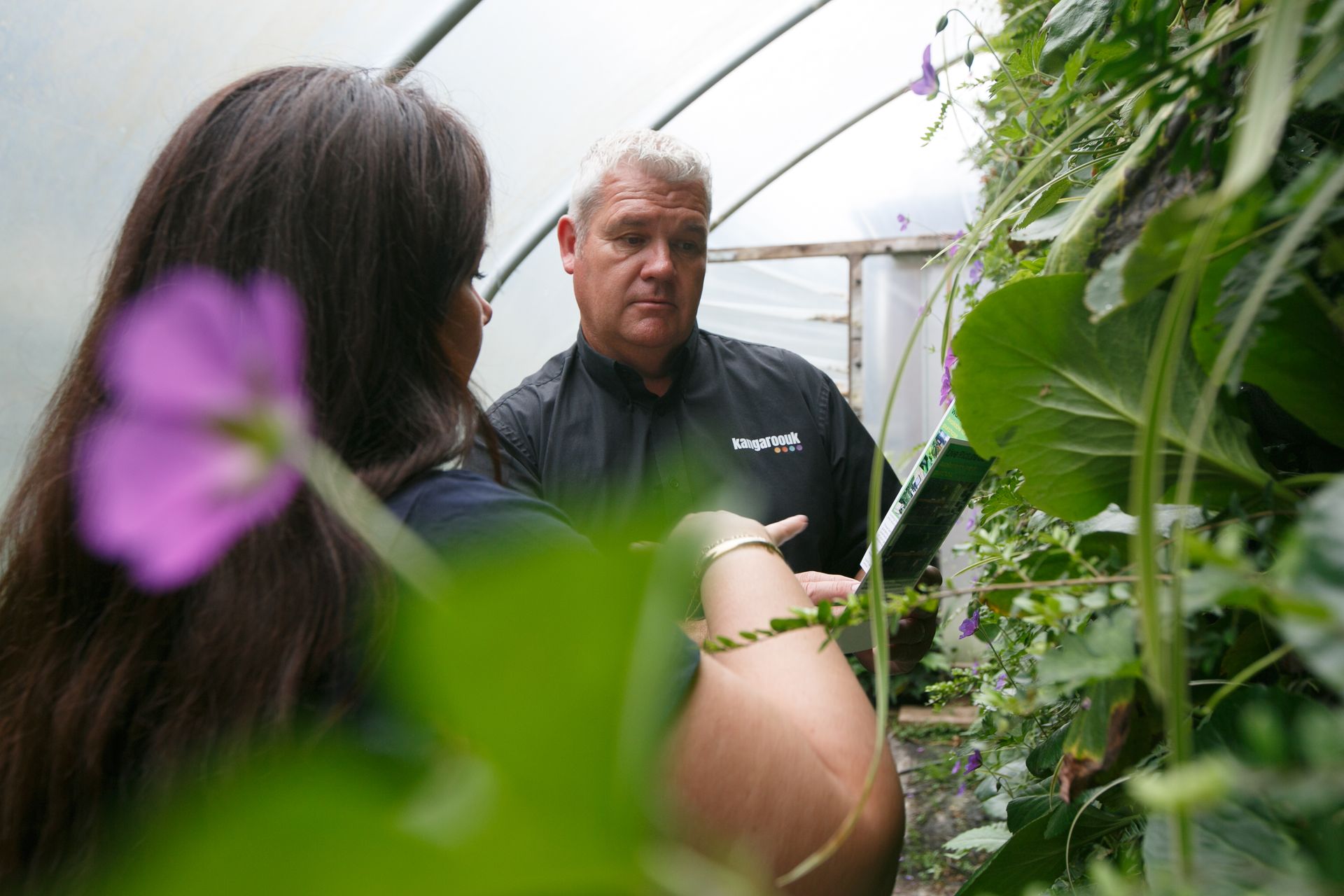 Two people in a greenhouse examine a vine-covered wall, one holding a green clipboard.