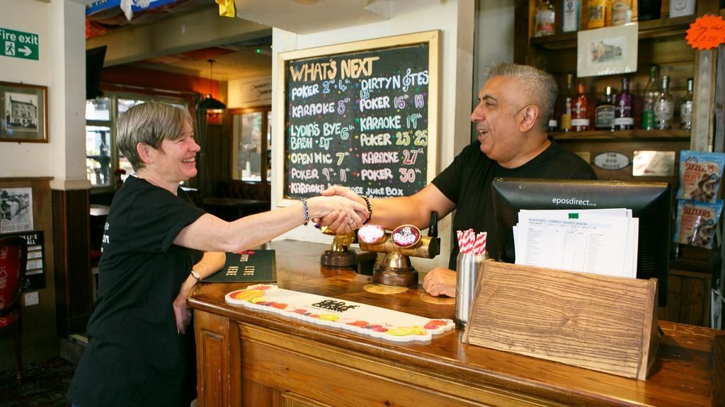 A person shakes hands with a bartender across a pub counter, with a chalkboard menu of drinks visible in the background.